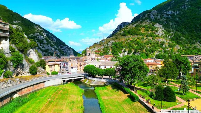 Picture-perfect aerial push-in shot of the Pioraco town center, drone flying above the Potenza River, approaching trees, the bridge, road, cars, tightly knit buildings, belltowers, majestic Sibillini