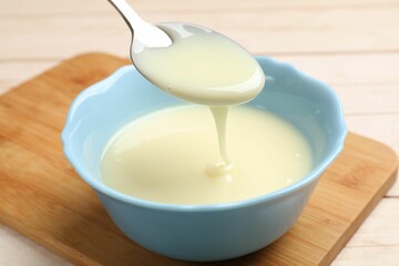 Condensed milk flowing down from spoon into bowl on wooden table, closeup