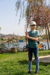 Young boy holding bow outdoors in park on sunny day, standing near picturesque lake and surrounded by spring trees. Concept of outdoor activity, childhood hobbies, and traditional archery.