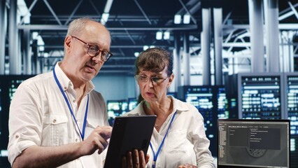 Older coworkers in data center, solving tasks on tablet while running scripts on laptop terminal....