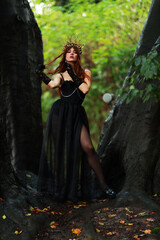 Naklejka premium Full length portrait of young brunette woman in vintage black dress and crown at old trees