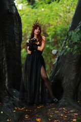Naklejka premium Full length portrait of young brunette woman in vintage black dress and crown at old trees