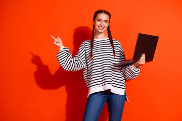 Cheerful woman in pullover holding laptop and pointing on vibrant orange background, highlighting...