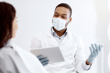 Young Black Dentist Doctor With Digital Tablet In Hands Consulting Patient In Clinic, African American Stomatologist Explaining Teeth Treatment Process To Customer During Check Up In Dental Cabinet