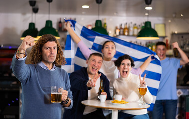 Group of emotive friends supporters cheering for favorite team with flag of Greece while watching match on TV in pub