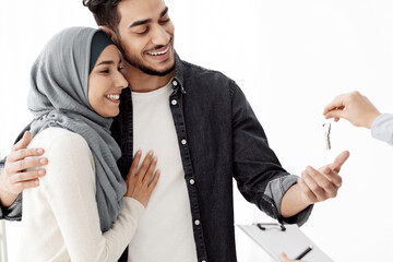 Happy young muslim family husband and wife in hijab relocating or buying house, unrecognizable landlord or real eastete agant giving cheerful middle-eastern couple key from apartment, copy space
