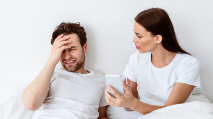 Jealous Wife Showing Cheating Husband His Phone With Message From Lover Demanding Explanation Suspecting Affair Sitting In Bedroom Indoor. Marital Issues, Jealousy In Relationship. Selective Focus