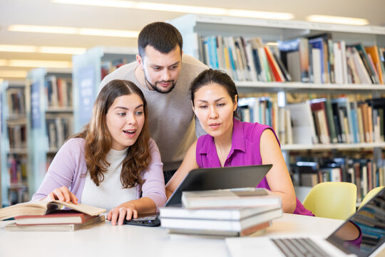 Friendly group of students studying in the university library is preparing for classes on a laptop, discussing current issues - Powered by Adobe