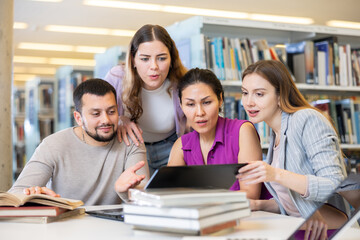 Fototapeta premium Cheerful young adult university students studying at library using books and laptop, talking and smiling while working on group project