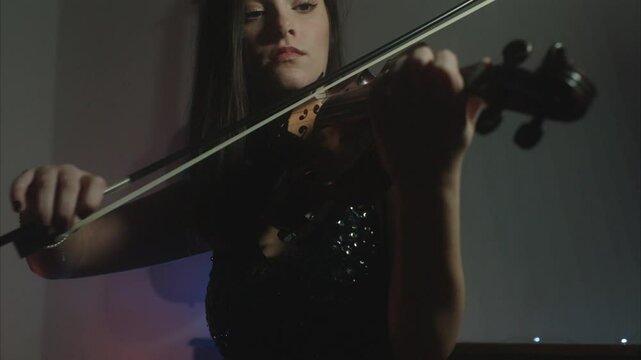 Beautiful young female violin player wearing a glitter black evening dress elegantly playing fiddle in a dark and hazy indoor environment 