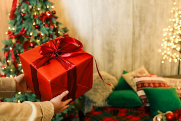 Person unwrapping a festive gift box with a red ribbon near a beautifully decorated Christmas tree. Cozy holiday atmosphere with pillows, candles, and glowing fairy lights in the background