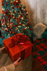 Person unwrapping a festive gift box with a red ribbon near a beautifully decorated Christmas tree. Cozy holiday atmosphere with pillows, candles, and glowing fairy lights in the background