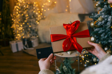 A woman holding a gift box with red ribbon and a credit card, shopping online using a laptop, with a glowing Christmas tree in the background, creating a festive and cozy atmosphere