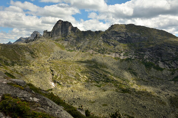 Fototapeta premium Steep rocky slopes of a high cliff with pointed peaks covered with grass in places under a cloudy summer sky.