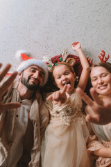 A family celebrates Christmas together. Father, mother and little daughter in festive costumes lie on the floor and wave, laugh and tickle their daughter. The little girl has fun with her parents.