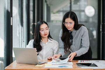 Two businesswomen working together in a contemporary office, analyzing documents and using a laptop for strategic planning.