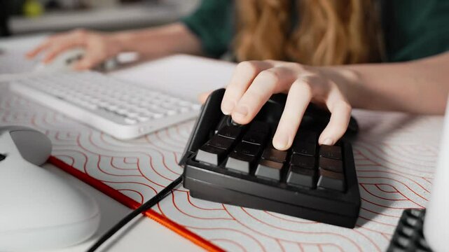 Close-up on hands of girl gamer playing in video game using keyboard and keypad, on table in the frame are gamer accessories. Young woman in cozy home office