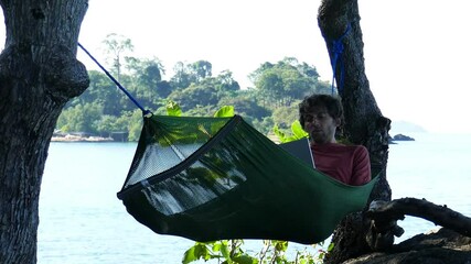 a man works at a laptop while lying in a hammock on a wild seashore
