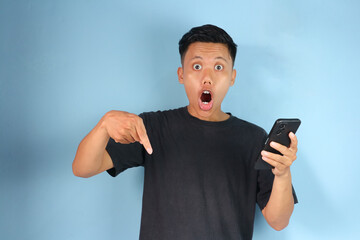Shocked face Asian man pointing down while holding mobile phone 