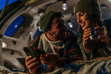 A happy couple staying warm with hot drinks in their truck on a frosty evening and using a smartphone