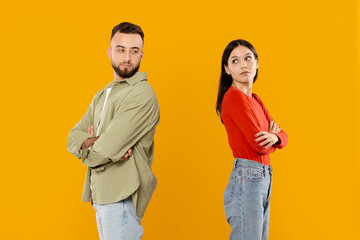 Fototapeta premium A young married couple stands back to back, both with crossed arms, showcasing contrasting emotions. The vibrant yellow background highlights their connection and tension in a playful way.