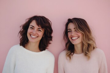 Two women in cozy sweaters share joyful smiles, standing together against a soft pink wall, radiating happiness and friendship.