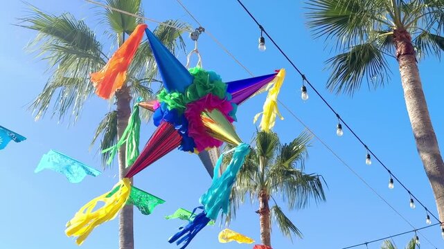 A brightly colored pi&ntilde;ata hangs suspended against a backdrop of swaying palm trees and a cloudless sky. Festive paper decorations, fluttering in the breeze, add to the celebratory atmosphere.