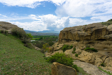 Gorge between green grass field and sandstone cliffs. Mountains, hills, blue sky with clouds in the background