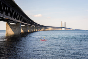 Two kayakers in a tandem sea kayak pass under the Oresund bridge, a railway and motorway cable-stayed bridge across the Oresund strait between Denmark and Sweden.