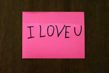 A Pink Sticky Note with a Handwritten Message I Love You on a Dark Wooden Surface