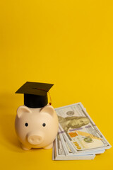 Vertical image of piggy bank with a mortarboard next to a wad of hundred dollar bills on an isolated yellow background, concept of saving for education
