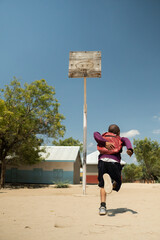A child running towards a basketball hoop in a rural schoolyard, carrying a red backpack. The scene features a sunny day with a clear blue sky, trees, and simple school buildings in the background.  © Roberto Lomba