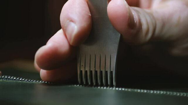 Worker stabs leather sheet with chisel to sew zipper line in shop closeup. Craftsman makes hole in genuine cowhide purse detail at table