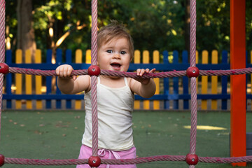 Fototapeta premium Cute little girl in pink top on children's playground. Happy child standing and holding on to wicker net. Concept of motherhood, happy childhood. High quality photo