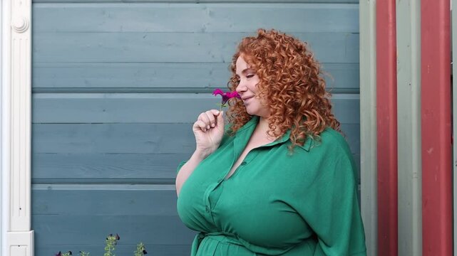 Plus Size Woman with Curly Red Hair in Green Clothing Holds Flower Against Vibrant Blue Wall Background, Beauty and Confidence Outdoors