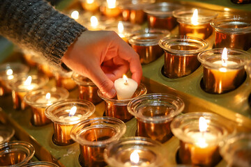 A close-up of a person's hands lighting a small candle among a row of glowing candles in a religious setting, symbolizing prayer, hope, and spirituality.