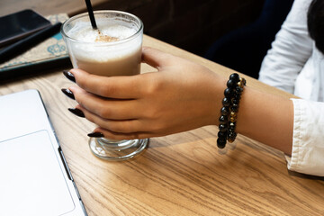 Transparent glass of aromatic cappuccino with foam in a woman's hand. Close-up. Side view. Office worker's hands. Unrecognizable girl holds a cup of coffee in front of a laptop.