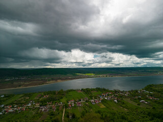 Summer storm clouds seen from a drone over a vast green forest. A village with houses gathered in a fairy tale landscape near a big lake