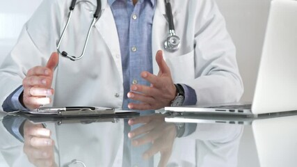 Male doctor wearing a blue shirt, stethoscope and lab coat is gesturing with hands while explaining a diagnosis to a patient across a glass desk with a laptop. Medicine concept