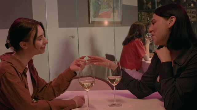 Two women meet in a cafe, sharing white wine and engaging in open flirtation. One tucks her hair behind her ear and rests her hand on her partner's, signaling mutual attraction.