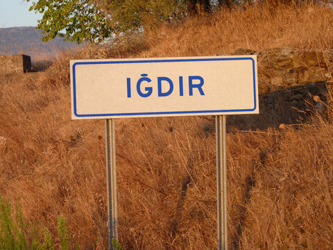 Blue 'Igdir' Sign on a Metal Pole: A Bold Contrast Against the Natural Landscape of Brown Grass and Soft Shadows, Capturing a Serene Afternoon in the Heart of the City.
