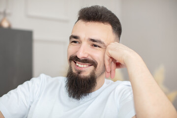 A man with a beard and a smile on his face. He is wearing a white shirt and leaning on his right hand.