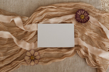 Blank white paper card mock up on a beige fabric background, ribbons and flowers. Aesthetic minimalistic business brand template, wedding invitation design, greeting card, copy space