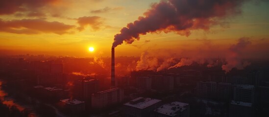 Fototapeta premium Smoke billowing from a tall chimney at sunset, casting a dark silhouette against the bright orange sky.