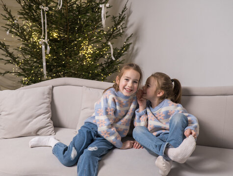 Two happy sisters, wearing matching pastel sweaters and jeans, sit on a light gray couch near a decorated Christmas tree. One whispers a secret to the other