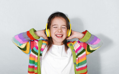 A joyful girl wearing bright yellow headphones and a colorful striped cardigan enjoys listening to...