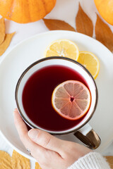 Female hand is holding cup of hibiscus tea with lemon on white marble table with autumn leaves and pumpkin. Top view. Woman is relaxing with homemade hot drink.