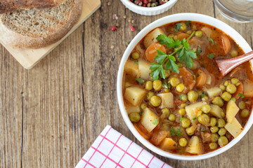 Bowl of vegetable stew with potatoes, green peas, and carrots on rustic wooden table. Top view. Meatless homemade meal.