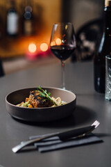 Gourmet dish in focus, featuring grilled meat with garnish in a ceramic bowl. Close-up composition with shallow depth of field, soft bokeh of candlelit background, and wine glass adding elegance.