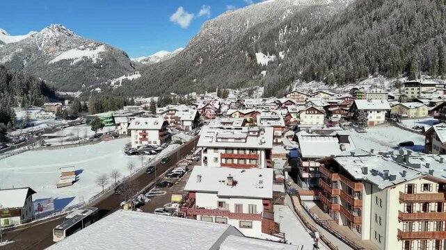 Canazei, Italy: Aerial view of ski resort in snowy Dolomites. Charming town landscape with chalets and hotels surrounded by snow-capped peaks, sunny day, clear blue sky
